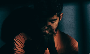 Image of incarcerated individual in dark area with hands together wearing and orange shirt