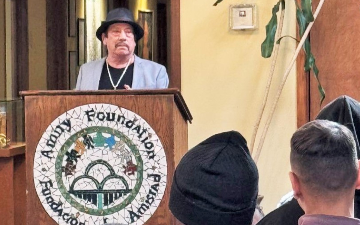 Danny Trejo visits the Male Community Reentry Program (MCRP) in Los Angeles. He's shown standing behind a lectern while people listen to him.