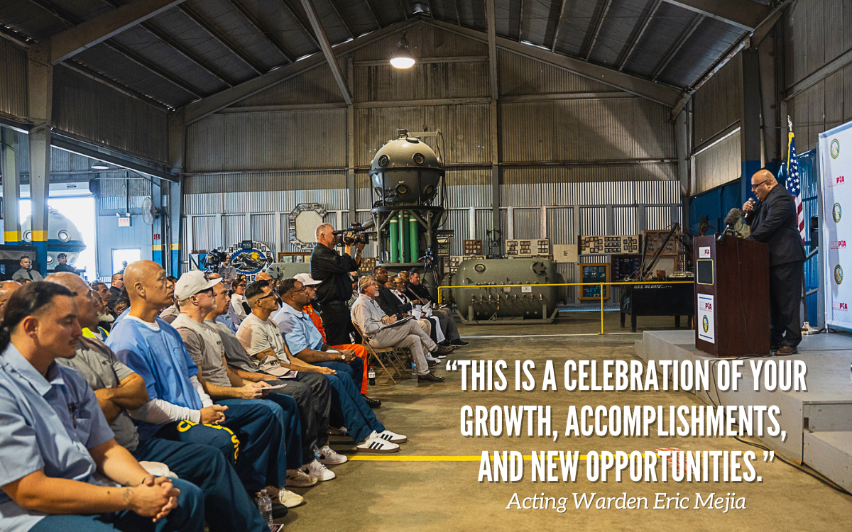 CALPIA dive center graduates with the CIM warden speaking at a lectern.