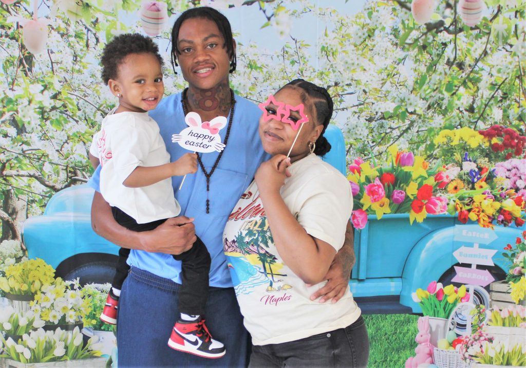 An incarcerated person holds his child while hugging a woman in front of an Easter/spring photo backdrop at Centinela State Prison.