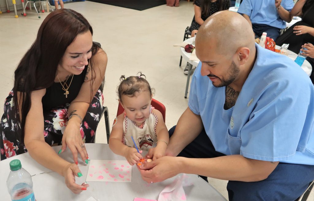 A young child plays with her incarcerated father while her mother smiles during Mother's Day weekend visiting at Calipatria State Prison.