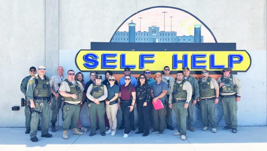 CSP-Corcoran staff pose for a group photo in front of a self-help mural.