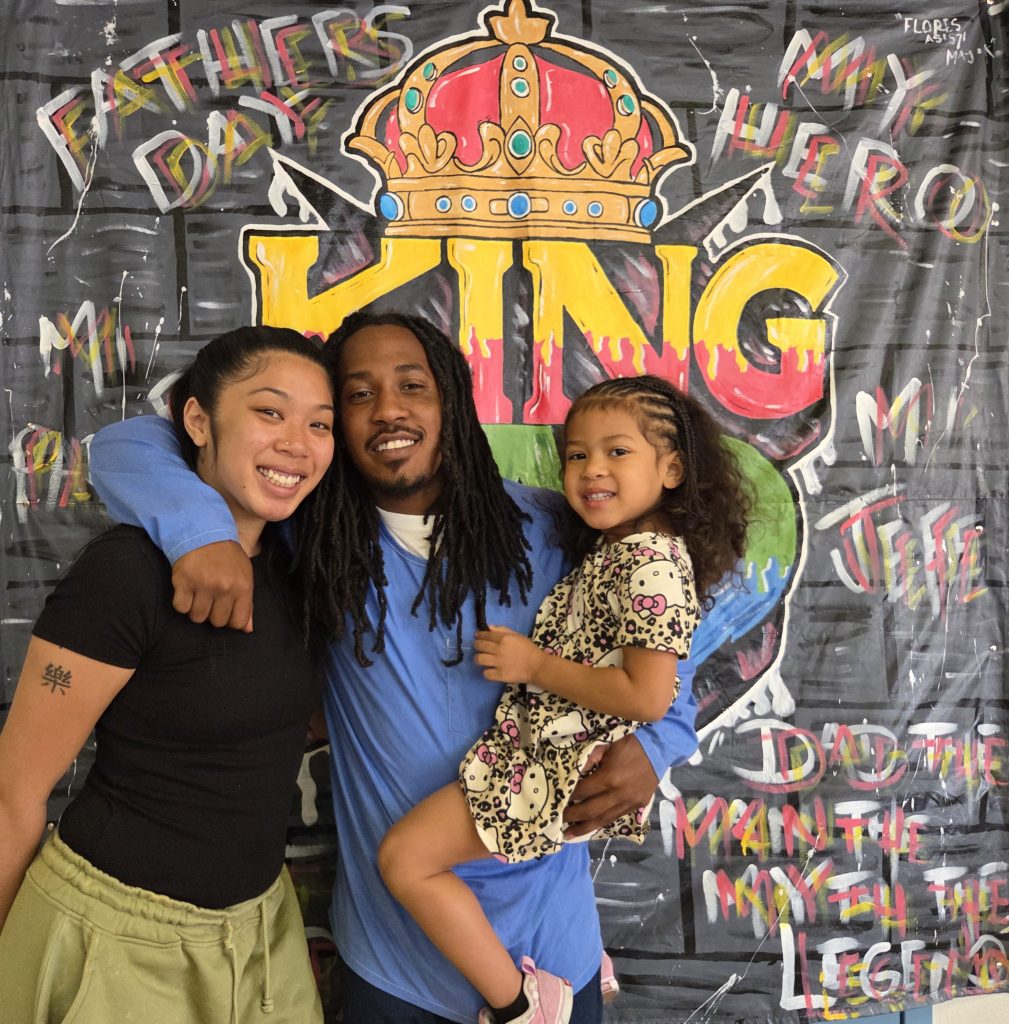 An incarcerated person hugs a woman while holding a little girl, standing in front of a King Dad banner with the words Father's Day and My Hero painted above it.