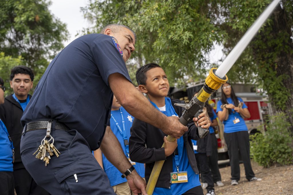 Staff member bends over to help young child hold fire hose at CIM.