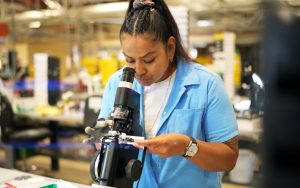 An incarcerated person is learning job skills at the Central California Women's Facility CALPIA Optical Program. She's seen looking through a piece of equipment to determine a lens prescription.