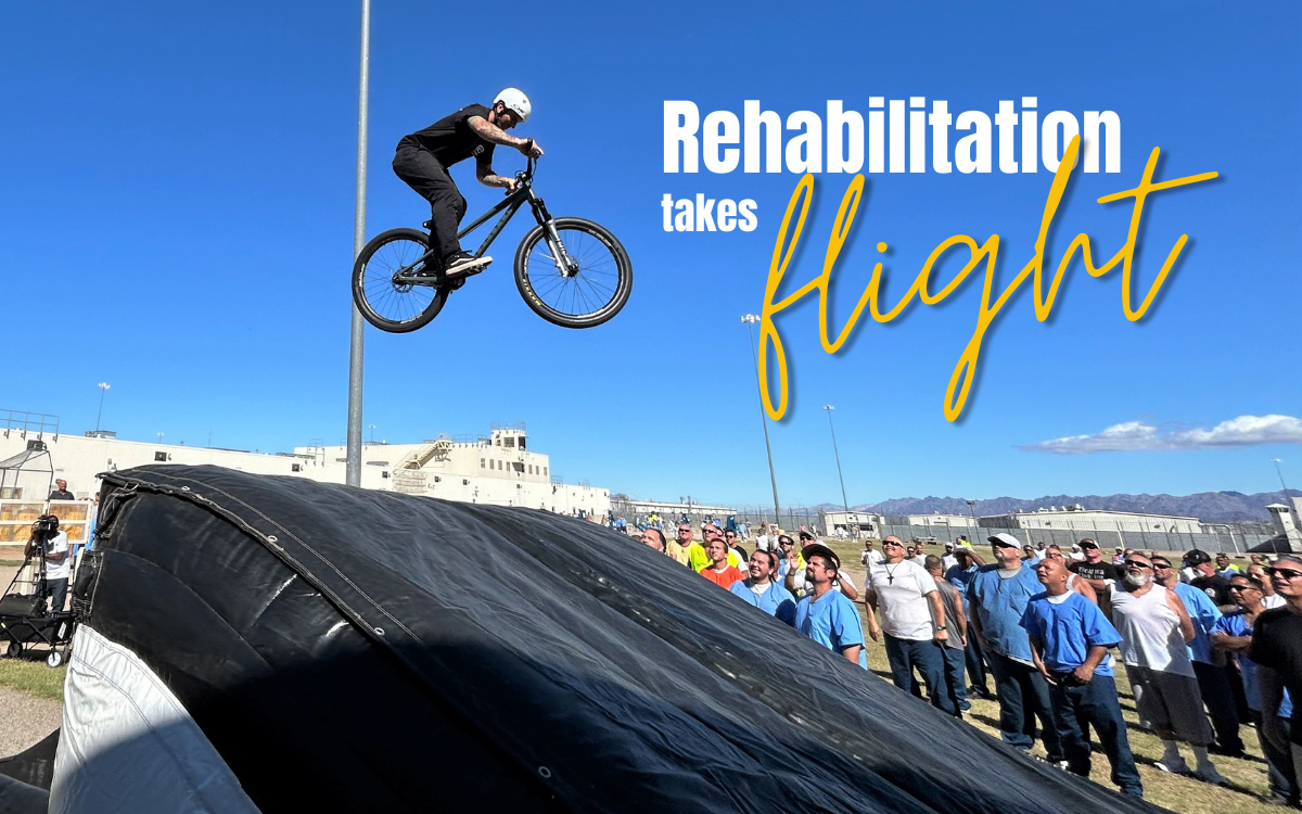 Ironwood State Prison rehabilitation event featured music, motorcycles and a BMX trick rider. The rider is seen in the air with the words "Rehabilitation takes flight" set against a blue sky while incarcerated people watch the BMX trick.