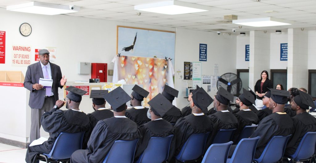A group of Peer Support Specialists dressed in graduation attire sit and listen to Acting Warden Andre Green speak.