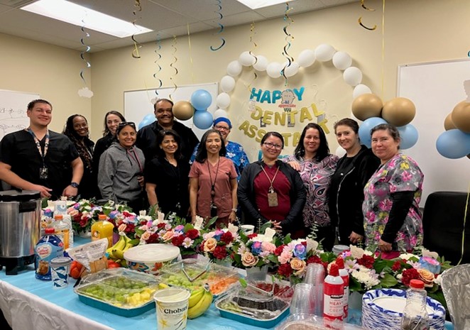 LAC dental assistants stand in front of table of food and flowers for dental assistant week celebration