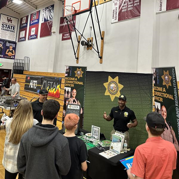 CDCR staff member stands behind table with recruitment materials and speaks to small group of high school students.