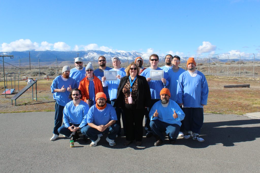A group of incarcerated graduates stand with female staff member holding graduation certificates