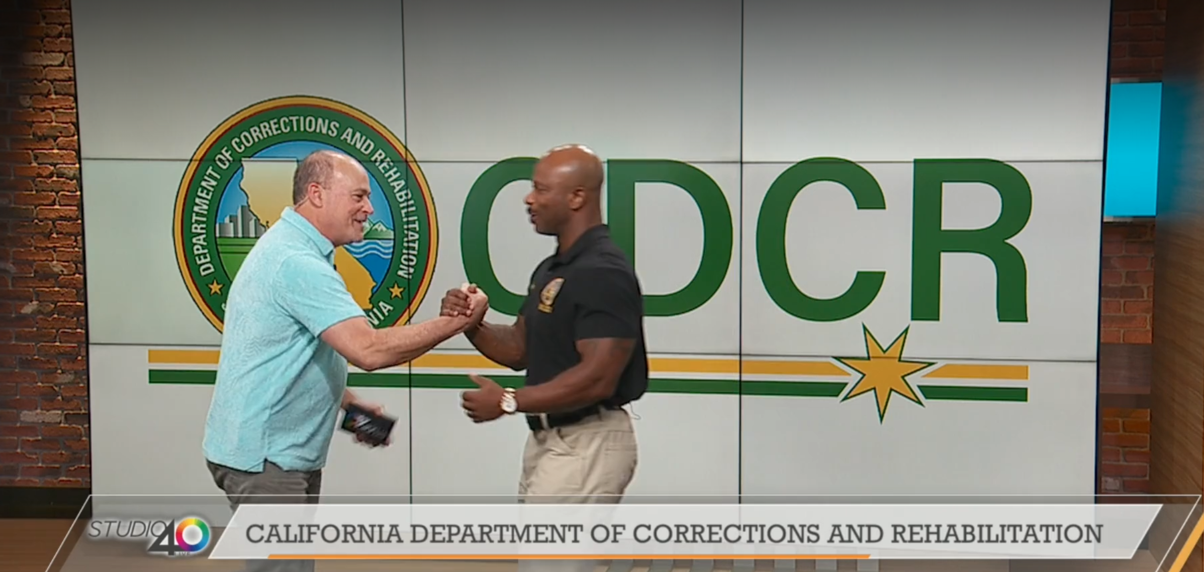 Two men in front of large CDCR backdrop lock hands together