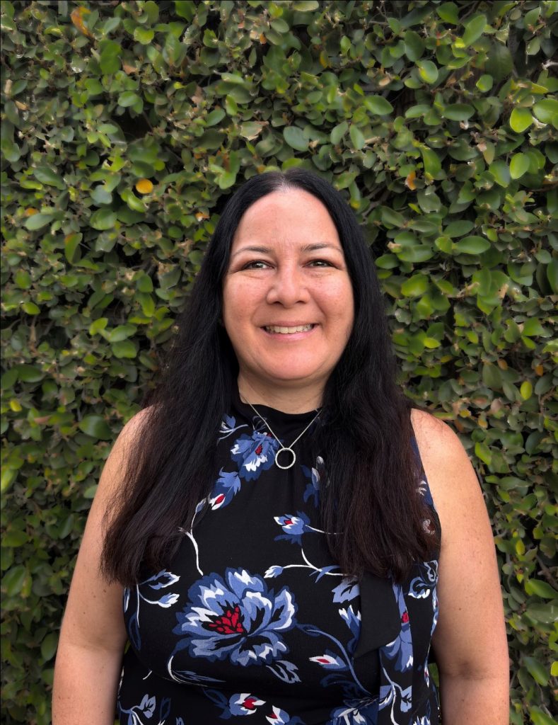 Woman smiling with dark hair standing in front of wall of greenery