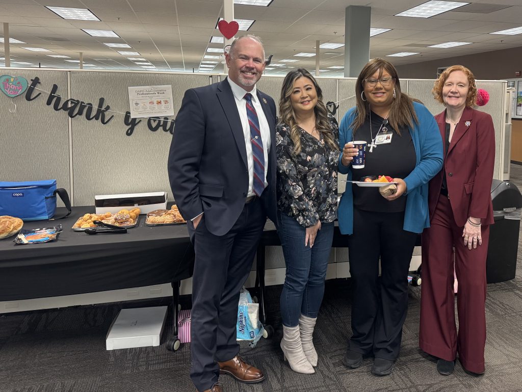 Four DAPO staff smile in front of table with refreshments and thank you sign for admin professionals week