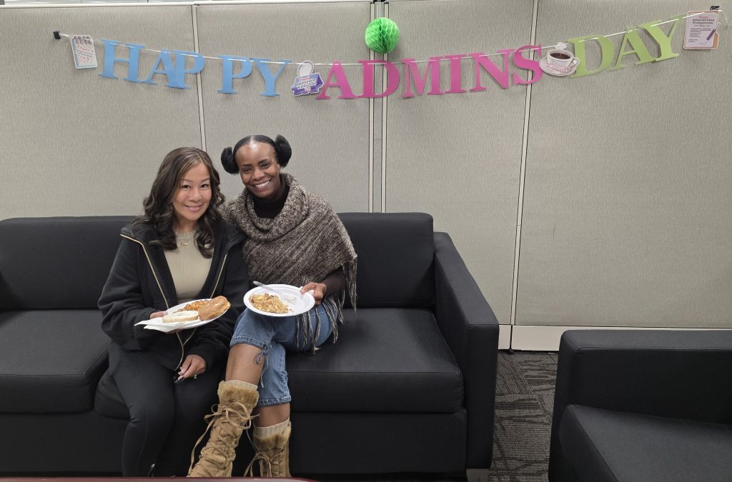 Two DAPO admin staff with plates of food and Happy Admins Day sign behind them