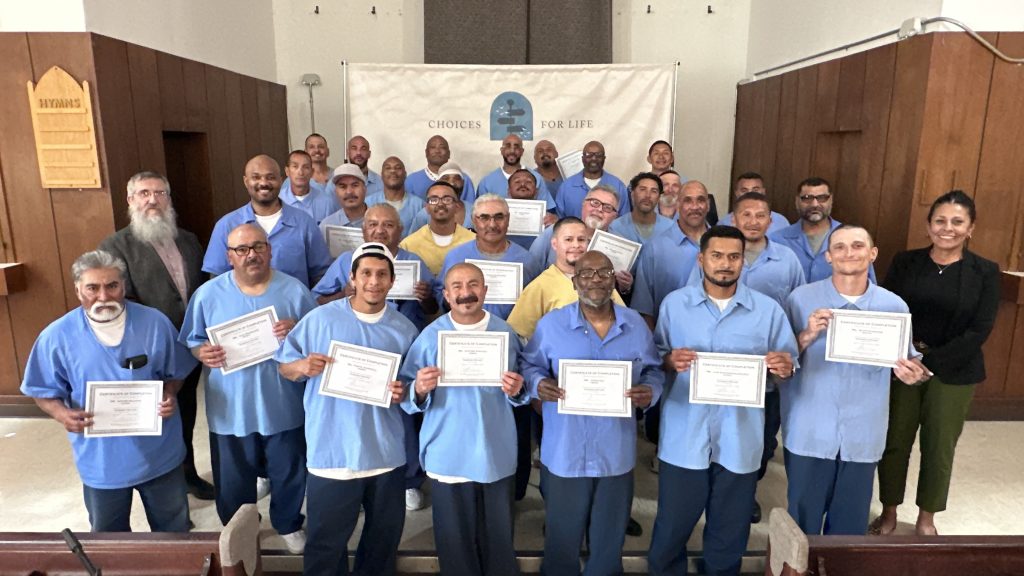 Multiple incarcerated individuals stand together holding graduation certificates