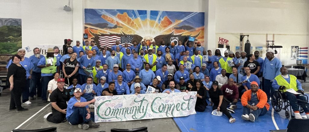 Large group of incarcerated individuals and staff gather at HDSP initiate justice event, some holding large sign with text "community connect"
