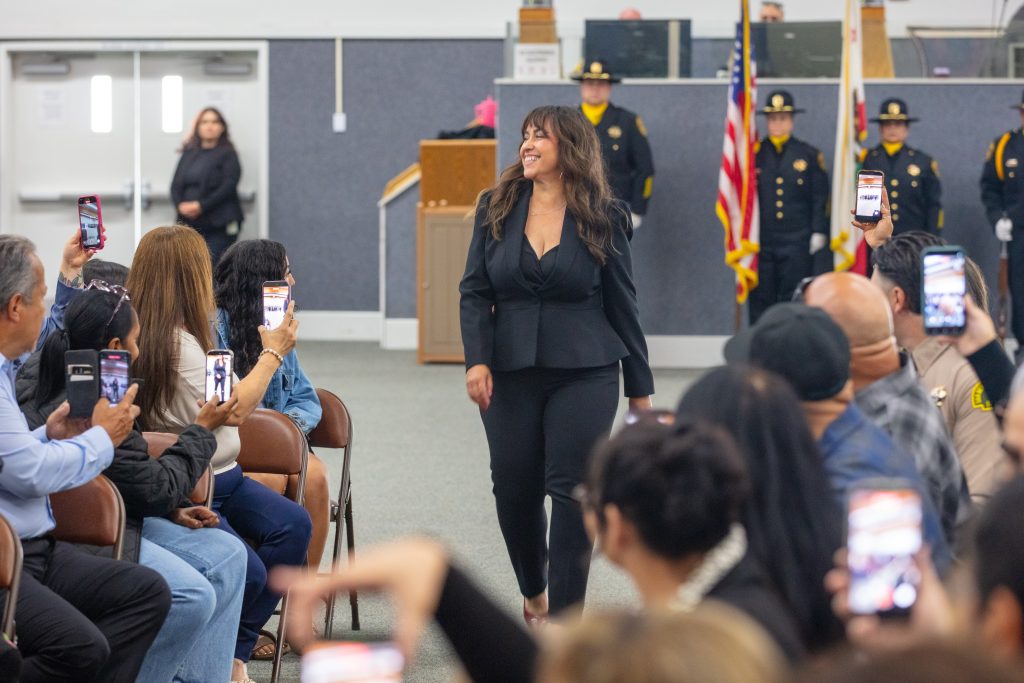 woman walking down walkway smiling with many audience members holding up phone recording, honor guard in background