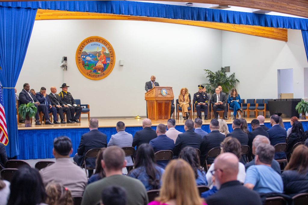 large room with many people in crowd facing speaker at podium and multiple participants in chairs on stage, large California seal on wall