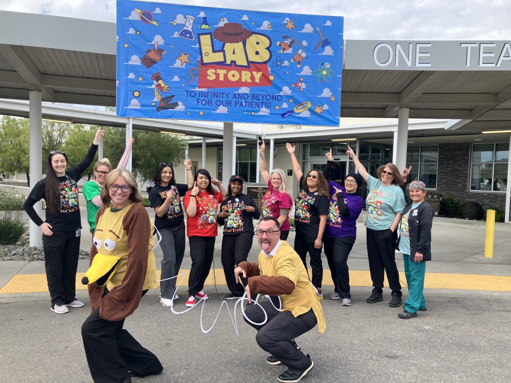 Med lab staff from CHCF pose in matching t-shirts with two staff members in front in "toy story" Slinky dog costume and large "Lab Story" banner in background.