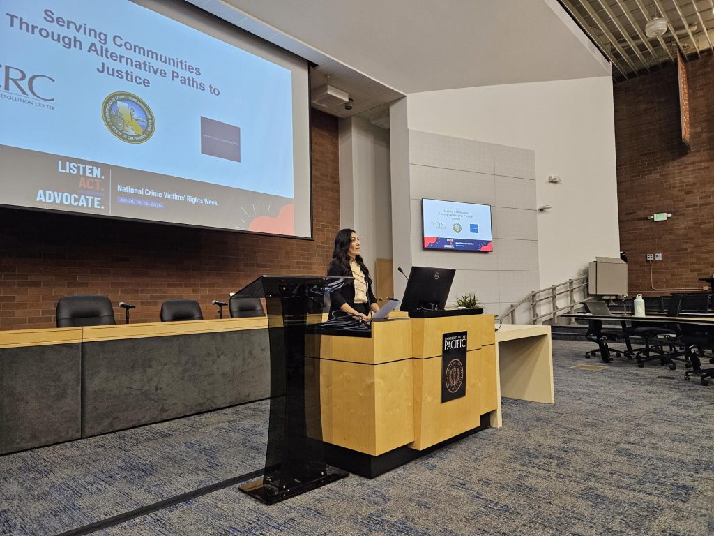 Woman presents at podium, large screen behind her with "serving communities through alternative paths to justice."