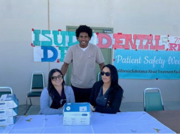 2 SATF staff sit at table, one stands behind them with ISUDT and Dental seen on sign behind them.,