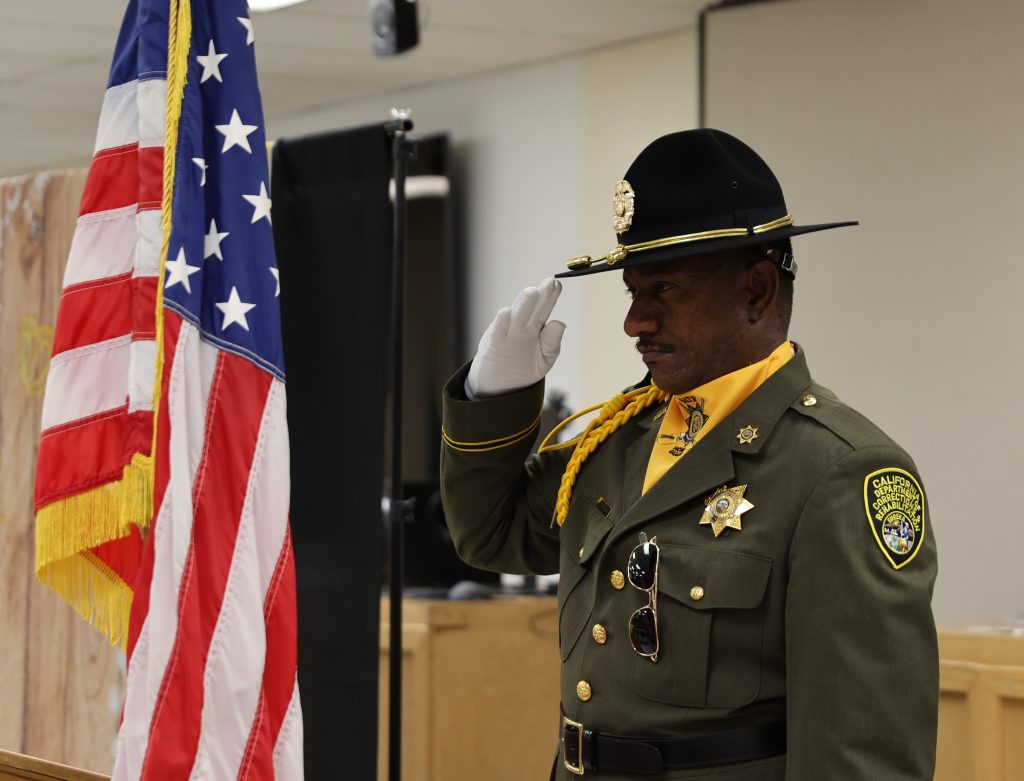 CDCR SATF honor guard member raises hand to hat with American flag in background