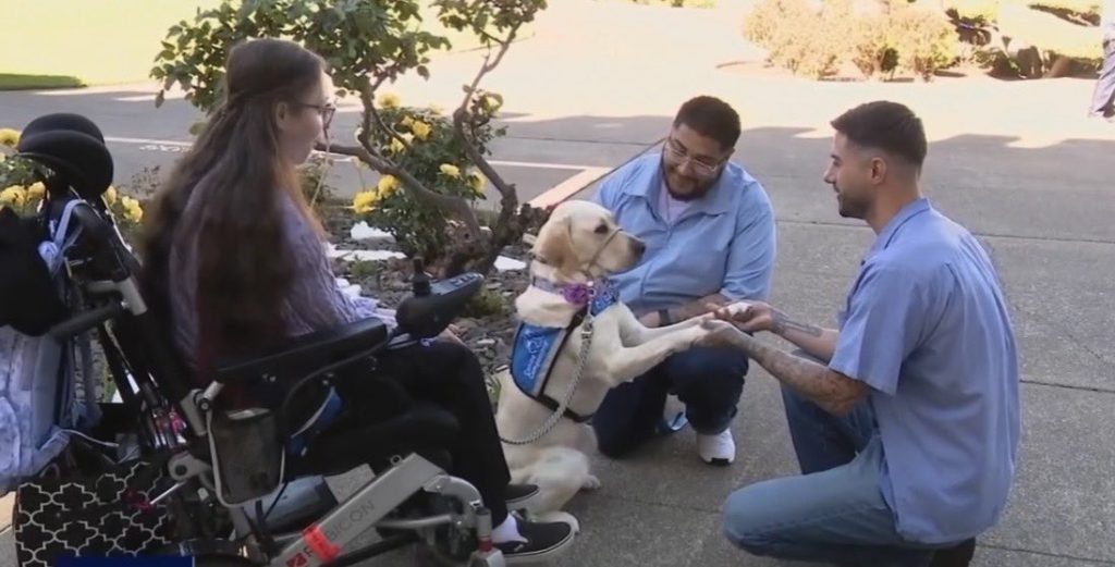 Two incarcerated individuals interact with service dog and woman in wheelchair