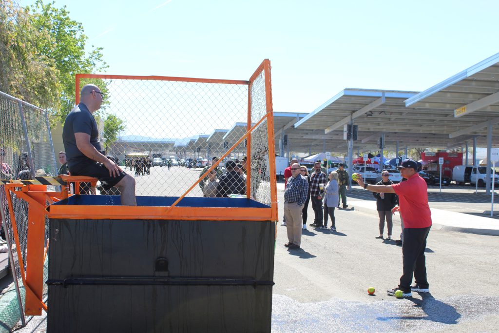 Man sits on dunk tank seat while another man gets ready to throw a ball. Staff standing in background.