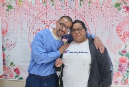 An incarcerated man hugs a woman who is holding a flower at Avenal State Prison during Mother's Day visiting.
