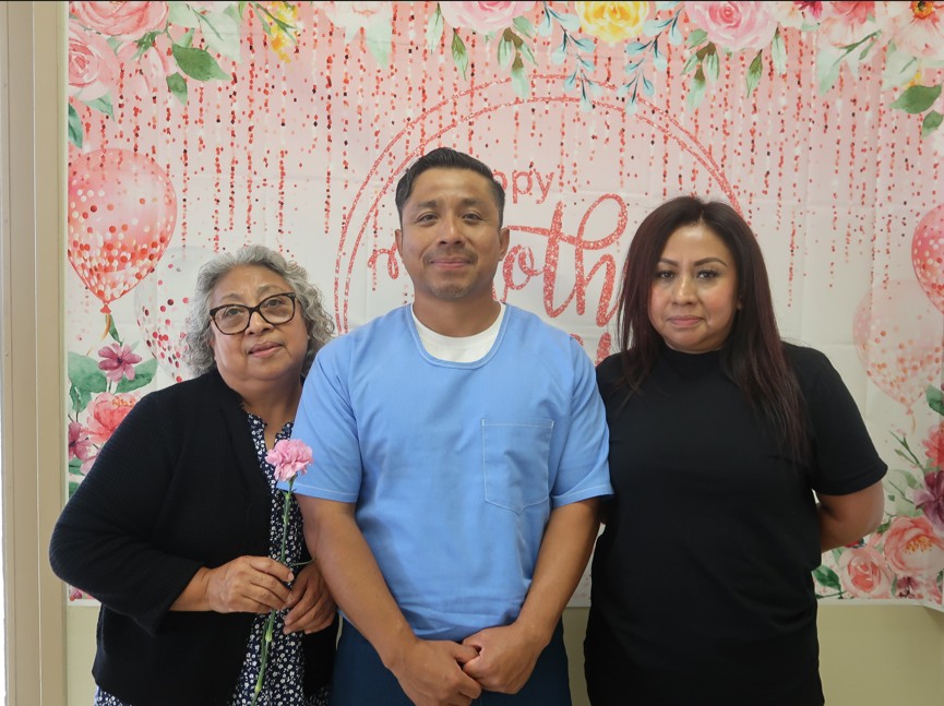 Two women flank an incarcerated man against a Mother's Day backdrop at Avenal State Prison.