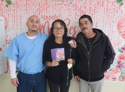 Three people, one of them incarcerated, at A woman holds a flower while standing beside an incarcerated man at Avenal State Prison during Mother's Day visiting.