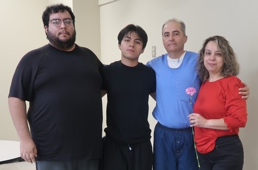 Four people, one of them incarcerated, at A woman holds a flower while standing beside an incarcerated man at Avenal State Prison during Mother's Day visiting.