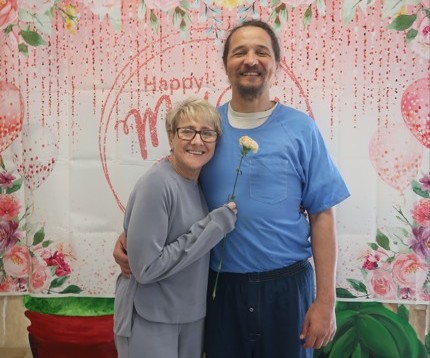 A woman holds a flower while standing beside an incarcerated man at Avenal State Prison during Mother's Day visiting.