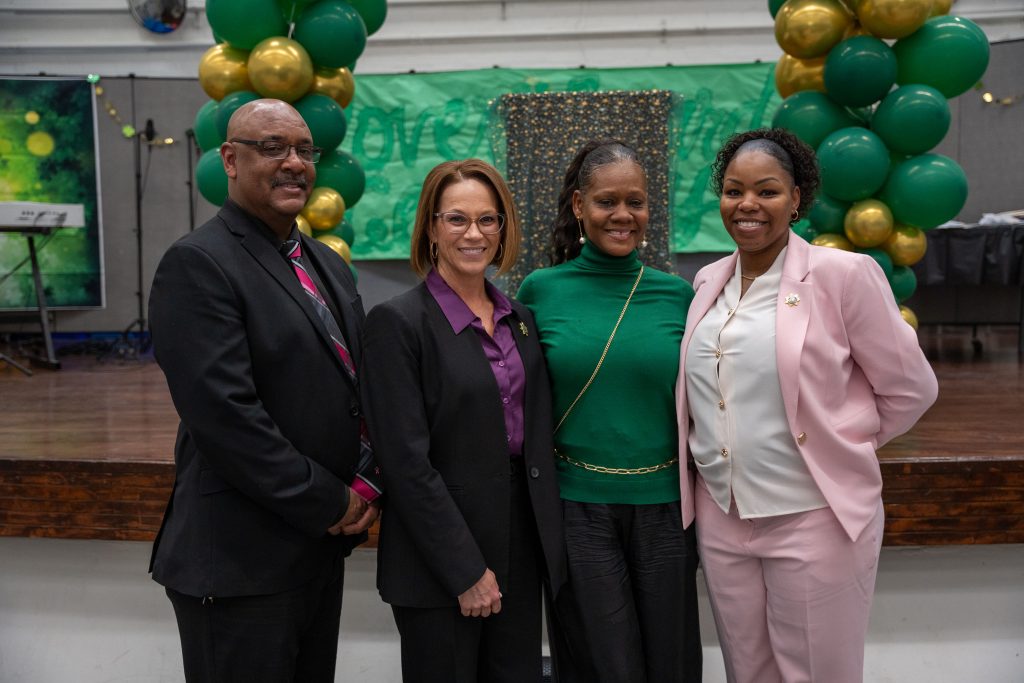 Four CIW staff stand in front of stage, green and gold balloons and large banner in background