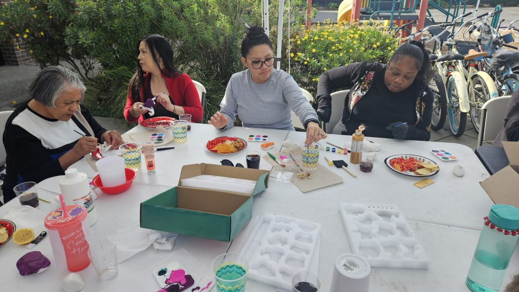 4 women sit at table at park, paint supplies and plates of food on table