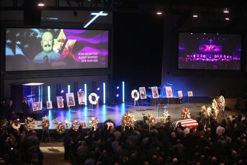 Wide overview photo of TCSO fallen officer Randy Hoppert funeral service, many people in audience, photos on stands, flower arrangements