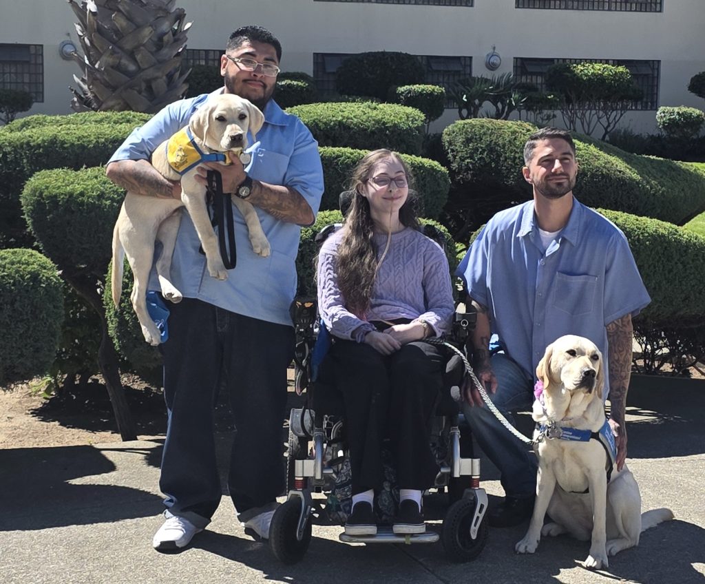 A dog with Canine Companions, two training handlers and a person being helped by the service dog.