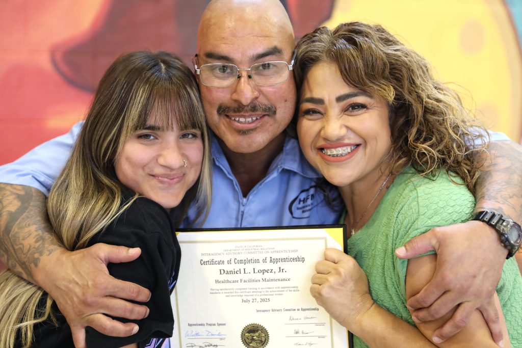 A graduate holds his certificate while his family hugs him at CSP-Solano's CALCTRA job certifications and apprenticeship graduation ceremony.