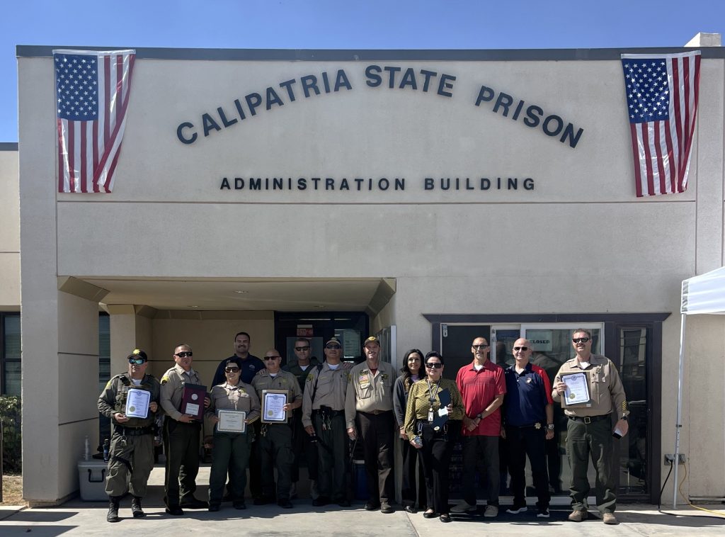 Group photo of people in front of the Calipatria State Prison Administration Building with flags hanging along the wall.