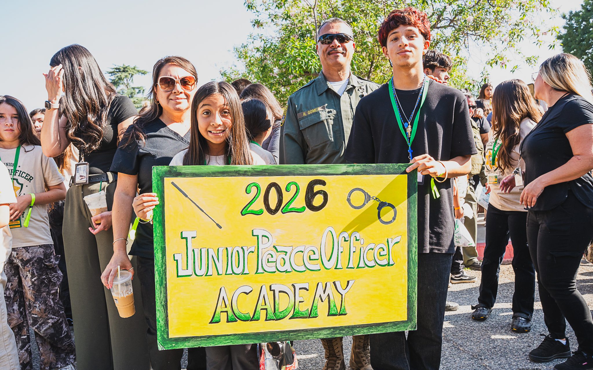 Two children hold sign with the words 2026 Junior Peace Officer Academy while adults and other children stand around them.