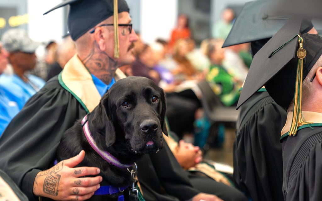 A dog in training sits with the students at Mule Creek State Prison for the CSU-Sacramento graduation.