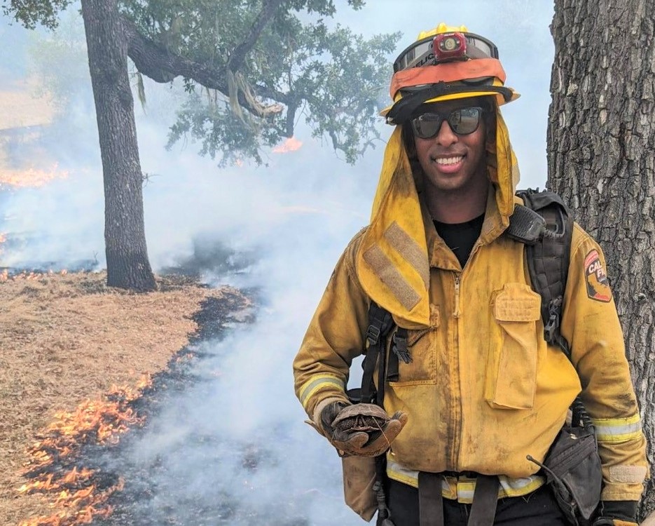 Michael Gebre in firefighter uniform with active tree and grass fire behind him