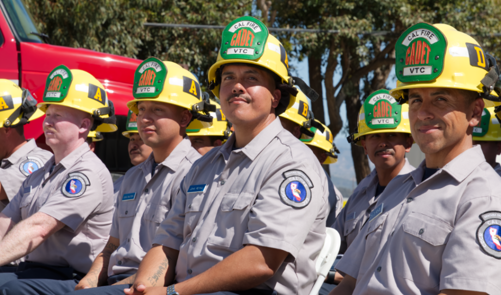 Cadets wearing Ventura Training Center uniforms and yellow CAL FIRE hard hats watch the speakers at graduation.