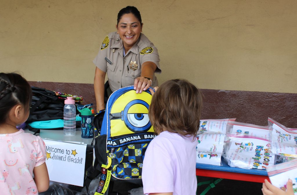 Uniformed officer smiles as she hands a minion backpack to a child.