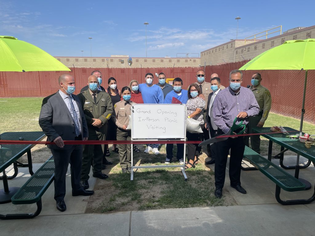 A group of masked people stand while a man cuts a ribbon with a pair of oversized scissors. A sign says "Grant opening in-person Picnic Visiting"