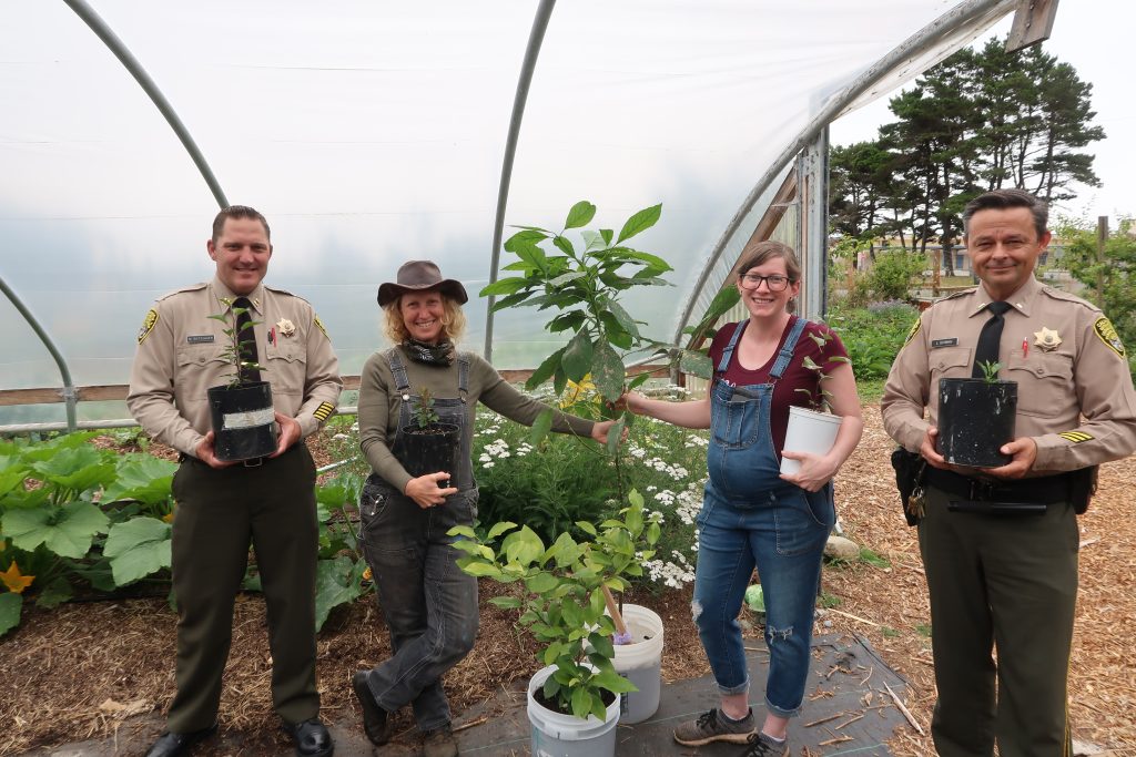 Two uniformed officers and two women in overalls stand in a greenhouse holding plants.