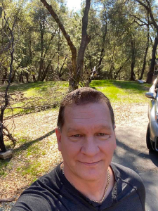 A man in a gray shirt takes a selfie in a wooded area