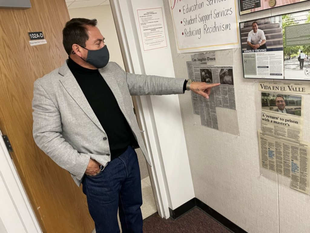 A man in jeans and a khaki blazer points to news articles taped to a wall
