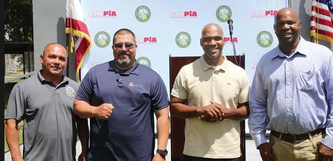 Four men stand in front of a stage with the PIA and CDCR logos
