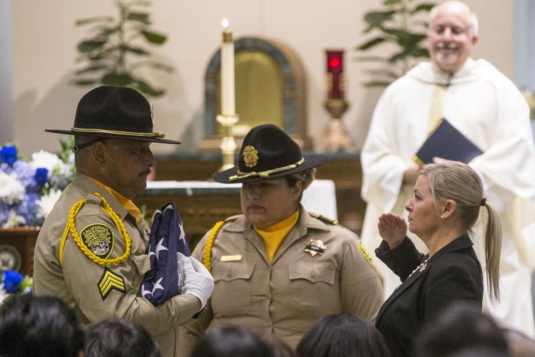 A woman in a black suit salutes two  corrrectional officers in uniform as one hands her a folded american flag. a priest looks on in the background.